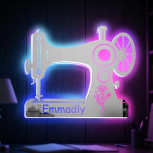 Puede incluir: Un letrero de pared plano en forma de máquina de coser, de color plateado, con retroiluminación LED azul y morada. La base muestra el nombre Emmadly en texto azul y un gráfico floral de líneas en rosa, montado con herrajes de color plateado.
