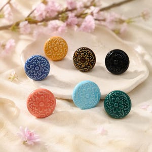 Puede incluir: Un conjunto de seis botones redondos de cerámica con patrones florales en relieve. Los botones tienen acabados brillantes en colores amarillo, marrón oscuro, negro, azul, coral, azul claro y verde oscuro, cada uno con un motivo floral central.