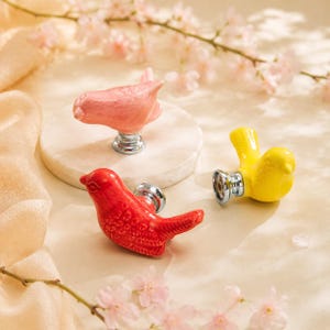 May include: Three bird-shaped cabinet knobs with a glossy finish. One knob is light pink, one is bright red, and one is vibrant yellow. Each bird figure is attached to a circular silver-toned metal base, designed for mounting onto furniture drawers.