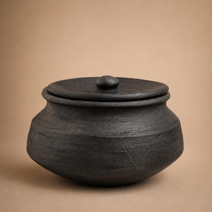 May include: A round, black clay pot with a matching lid. The exterior surface features a rough, textured finish with subtle, incised geometric triangle patterns around the midsection. The pot has a wide, curved body and a short, narrow neck.