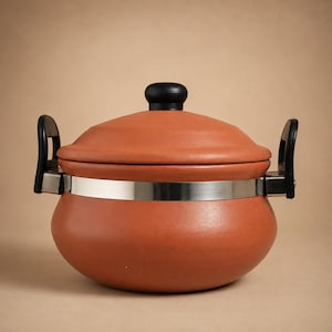 Pode incluir: Uma panela de terracota com corpo arredondado e tampa a condizer. A panela apresenta uma larga faixa metálica de cor prateada polida que rodeia o centro, ligada a duas pegas laterais de plástico preto e um botão preto na tampa.