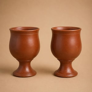 Puede incluir: Dos cálices de terracota a juego con un acabado mate y liso. Cada recipiente presenta un cuenco ancho y redondeado que se estrecha hacia un tallo delgado, apoyado sobre una base circular estable. La arcilla es de un color marrón rojizo.