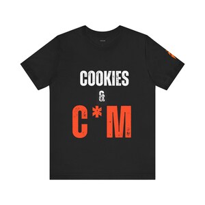 Puede incluir: Una camiseta negra de manga corta con el texto blanco COOKIES, un pequeño ampersand blanco y el texto naranja C*M con una textura desgastada. Un pequeño gráfico naranja es visible en el borde de la manga derecha y la camiseta tiene cuello redondo.