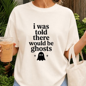 Puede incluir: Una camiseta de manga corta de color crema con el texto negro "i was told there would be ghosts" centrado en el pecho, acompañado de un pequeño icono de fantasma negro y dos estrellas de cuatro puntas situadas bajo el texto.