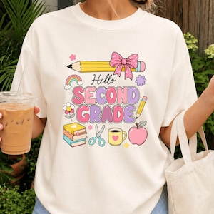 Puede incluir: Una camiseta de color crema con un gráfico que incluye el texto Hello SECOND GRADE. El diseño presenta un lápiz amarillo con un lazo rosa, un arcoíris, una pila de libros, tijeras, una taza amarilla con un corazón y una manzana rosa en tonos pastel.