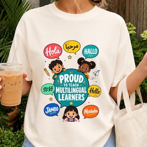 Puede incluir: Una camiseta de color crema con un gráfico de tres niños rodeando un gran círculo azul verdoso con el texto "PROUD TO TEACH MULTILINGUAL LEARNERS." El diseño incluye coloridos globos de texto con saludos en varios idiomas y pequeñas estrellas.