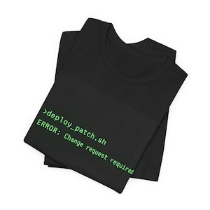 May include: A black t-shirt featuring green computer terminal text that reads >deploy_patch.sh followed by the error message ERROR: Change request required. The shirt has a crew neckline and short sleeves, presented in a folded, flat layout.
