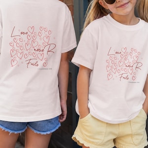 May include: A white short-sleeved t-shirt featuring a graphic print of numerous outlined hearts in shades of pink and red. The text "Love never Fails" is printed in a cursive script, accompanied by the reference "I Corinthians 13:8" below it.