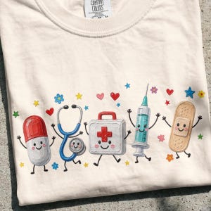 Può includere: Una t-shirt color crema con una stampa grafica di cinque oggetti medici antropomorfi: una capsula rossa e bianca, uno stetoscopio blu, un kit di pronto soccorso bianco con una croce rossa, una siringa turchese e un cerotto beige, tutti con volti.
