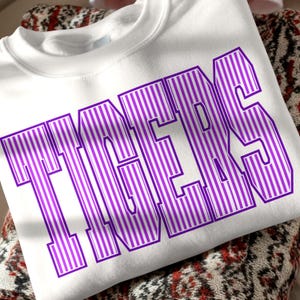 Puede incluir: Una sudadera blanca de cuello redondo con la palabra TIGERS impresa en letras grandes, negritas y de color morado. Cada letra contiene un patrón de rayas verticales en morado y blanco, creando un diseño gráfico distintivo en el pecho.