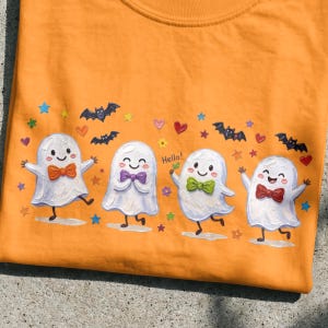 Pode incluir: Uma t-shirt laranja com uma impressão gráfica de quatro fantasmas brancos com rostos sorridentes, cada um usando uma gravata borboleta de cor diferente. O design inclui pequenos morcegos pretos, corações e estrelas coloridas e a palavra Hello.