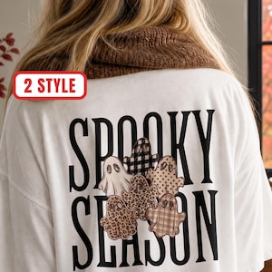 Peut inclure: Un t-shirt blanc avec le texte noir SPOOKY SEASON au dos. Le motif comprend cinq illustrations de fantômes avec divers motifs, notamment un tartan noir et blanc, un imprimé léopard marron et des pois beiges.