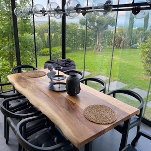 May include: A live-edge wooden dining table with a natural grain finish, paired with six black curved chairs featuring tufted seats. On the table sits a black three-tiered serving stand, a small white bust sculpture, and a black face-shaped vase.