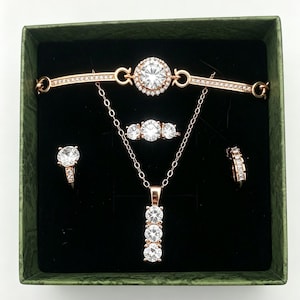 Könnte beinhalten: Ein Schmuckset aus Roségold, bestehend aus einem Armband mit einem runden Mittelstein, einem Ring mit drei Steinen, einer Halskette mit drei vertikalen runden Steinen und zwei Creolen, alle mit kleinen klaren Kristallen in einer grünen Box.