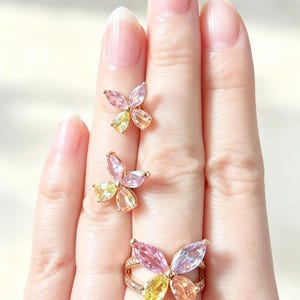 Peut inclure: Une bague et deux boucles d'oreilles assorties en métal doré, chacune arborant un motif de papillon composé de quatre pierres précieuses taille marquise dans des tons rose, jaune et orange. L'anneau est double et orné de petites pierres claires.