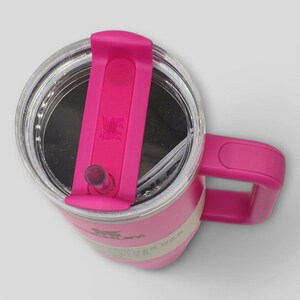 May include: A bright pink insulated tumbler with a clear lid and a matching pink handle. The lid features a rotating pink cover with a central straw opening and a small embossed logo. The side of the tumbler displays the Stanley brand name and H2.0 text.