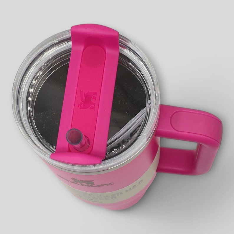 May include: A bright pink insulated tumbler with a clear lid and a matching pink handle. The lid features a rotating pink cover with a central straw opening and a small embossed logo. The side of the tumbler displays the Stanley brand name and H2.0 text.