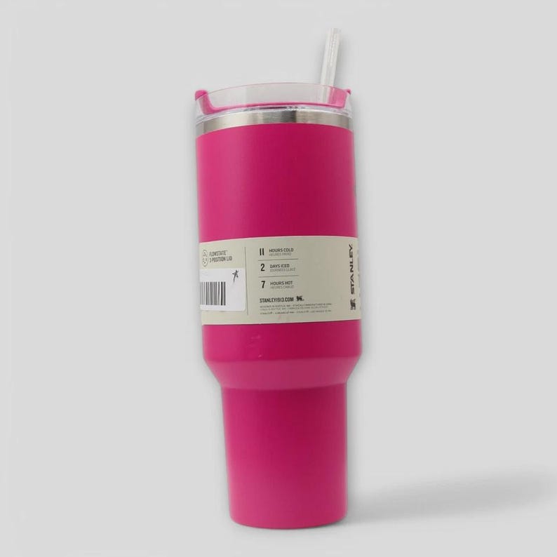 May include: A bright pink insulated tumbler with a clear lid and a white straw. A rectangular product label is wrapped around the center, displaying text about temperature retention times, a barcode, and the Stanley brand name in black ink.