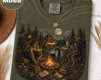 May include: An olive green t-shirt featuring a graphic print of a campfire scene in a forest with a sasquatch, an owl, and a deer-like creature reading books. The shirt has a crew neck and a Comfort Colors brand label printed on the inside neck.