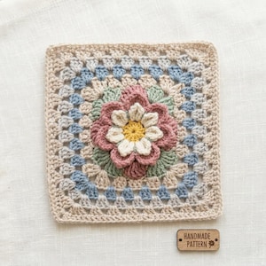 May include: A square crochet granny square featuring a central three-dimensional flower with yellow, cream, and dusty rose petals. The surrounding border incorporates sage green, light blue, and beige yarn in a classic stitch pattern.