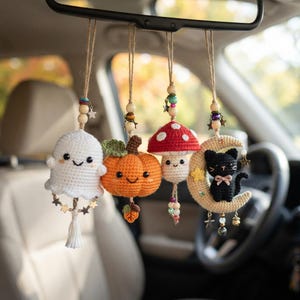 Könnte beinhalten: Vier hängende Häkelornamente für den Rückspiegel im Auto, bestehend aus einem weißen Geist, einem orangefarbenen Kürbis, einem rot-weißen Pilz und einer schwarzen Katze auf einer beigen Mondsichel, verziert mit Holzperlen und kleinen Metallsternen.