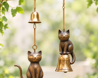 Könnte beinhalten: Zwei hängende Windspiele aus bronzefarbenem Metall. Eines zeigt eine Katzenfigur, die über einer Glocke sitzt, während das andere eine Glocke zeigt, die über einer Katze hängt. Beide Katzen haben geschlossene Augen und eine texturierte Oberfläche.