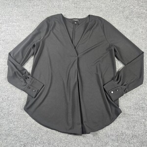 May include: A solid black long-sleeve blouse featuring a deep V-neckline, a central vertical pleat, and cuffs on each sleeve secured with two small black buttons. The garment has a curved hemline and a smooth, lightweight fabric construction.