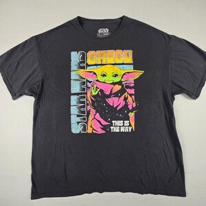 May include: A black short-sleeve t-shirt featuring a graphic of Grogu from Star Wars. The print includes the text STAR WARS, GROGU, and THIS IS THE WAY in vibrant neon colors against a dark background with a starry pattern and pink accents.