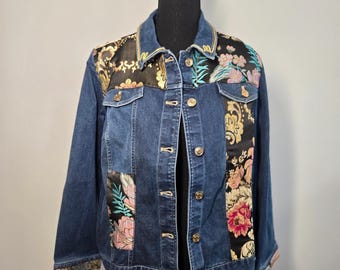 May include: A blue denim jacket featuring black fabric panels with gold baroque patterns and colorful floral embroidery. The jacket includes a classic collar, long sleeves, two chest pockets with flaps, and a front button closure with metallic buttons.