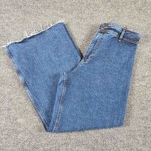 May include: Medium-wash blue denim jeans featuring a high-rise waist, a button and zipper fly closure, belt loops, and wide-leg openings. The bottom hems of the pant legs are finished with a raw, frayed edge detail, showcasing loose white threads.
