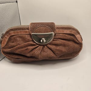 May include: A brown clutch bag with a textured snakeskin-like pattern, featuring a central silver-toned metal clasp with the engraved text 'botkier'. The bag has a rounded, pleated design with smooth leather trim along the top and zipper closure.