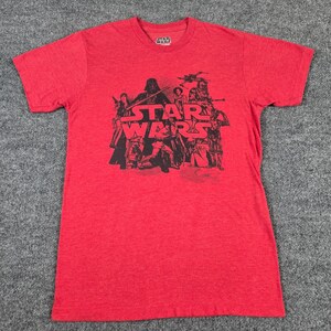 May include: A red short-sleeved t-shirt featuring a black graphic print of Star Wars characters including Darth Vader, Luke Skywalker, and C-3PO. The center of the chest displays the Star Wars logo in a bold, black font over the character illustration.