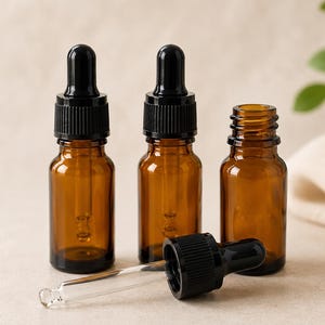 Puede incluir: Tres frascos cuentagotas de vidrio ámbar con tapas negras. Dos frascos están en posición vertical con sus pipetas insertadas, mientras que un frasco está abierto sin tapa. Una tapa cuentagotas negra con pipeta yace horizontalmente.