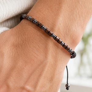 Pode incluir: Uma pulseira de contas com alternância de pequenas contas cilíndricas de madeira castanho-escura e contas espaçadoras cinzentas metálicas. A pulseira é feita num fio preto fino com um fecho de nó deslizante ajustável e duas pontas.