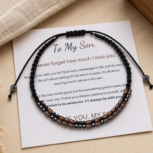 Könnte beinhalten: Ein schwarzes Kordelarmband mit abwechselnd einfarbig schwarzen und braun gemusterten zylindrischen Perlen sowie einem verstellbaren Schiebeknotenverschluss. Das Armband liegt auf einer weißen Karte mit dem Text To My Son, never forget how much I love you.