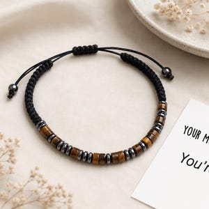 Può includere: Un bracciale con perline cilindriche alternate in occhio di tigre marrone ed ematite grigio metallizzato su un cordino nero. Il bracciale presenta una chiusura a nodo scorsoio intrecciato nero con due piccole perline grigio metallizzato alle estremità.
