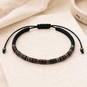 May include: A bracelet featuring alternating dark brown cylindrical wooden beads and small metallic gray disc beads strung on a black braided cord. The adjustable closure consists of a sliding knot mechanism with two black cord ends finished with beads.