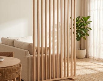 Puede incluir: Un separador de ambientes alto y rectangular fabricado en madera de color claro. La estructura presenta una serie de listones verticales dispuestos en un patrón paralelo, sostenidos por una base horizontal sobre el suelo.