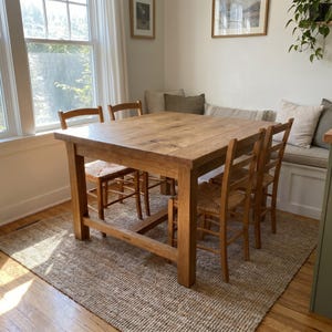 May include: A square wooden dining table with four legs and a natural light brown finish, accompanied by four wooden chairs featuring woven seats. The table has a smooth, flat surface and a sturdy frame, positioned on a textured woven area rug.