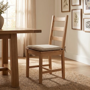 Puede incluir: Una silla de comedor de madera de color marrón claro con diseño de respaldo de escalera y asiento cuadrado. La silla cuenta con un cojín de tela beige sujeto con pequeñas cintas en las esquinas traseras. La estructura es de madera natural lisa.