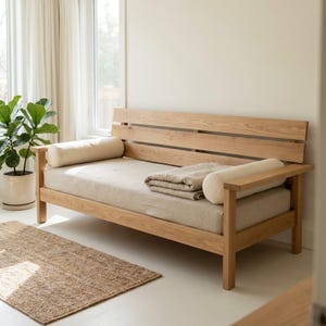 May include: A light oak wood daybed featuring a slatted backrest, a beige upholstered seat cushion, two cylindrical beige bolster pillows at each end, and a folded beige textile blanket placed on the center of the seat cushion.