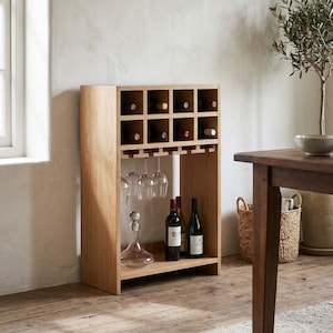 May include: A light oak wood wine rack featuring eight individual bottle storage cubbies at the top, a hanging stemware rack for wine glasses, and an open lower shelf holding a glass decanter and two wine bottles with dark labels.