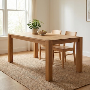 Puede incluir: Una mesa de comedor rectangular de madera marrón claro con cuatro patas gruesas, acompañada de dos sillas de madera a juego. Una pequeña planta verde en maceta y un cuenco blanco con varias frutas verdes y rojas descansan sobre la mesa.