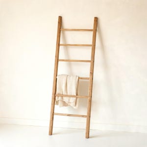 May include: A tall, light brown wooden ladder with five horizontal rungs. A folded, cream-coloured textile is draped over the third rung from the top. The ladder is constructed from smooth, natural wood with a visible grain and a simple, vertical design.