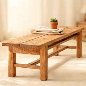 May include: A rectangular wooden bench with a natural grain finish, featuring four sturdy legs and a horizontal support brace. A small terracotta pot containing a green succulent and a folded beige fabric cloth rest on the bench surface.
