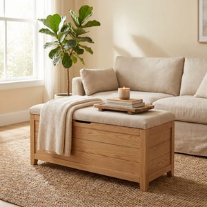Könnte beinhalten: Eine rechteckige Aufbewahrungsbank aus hellem Holz mit horizontaler Paneeloptik und einer gepolsterten Sitzfläche aus beigem Stoff. Eine cremefarbene Strickdecke liegt über einer Seite der Bank, die mit einem Klappdeckel ausgestattet ist.