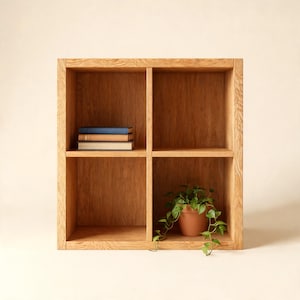 Puede incluir: Una estantería cuadrada de madera con cuatro compartimentos abiertos. El estante superior izquierdo contiene una pila de tres libros con cubiertas azul, beige y blanca. El estante inferior derecho contiene una planta verde en una maceta de terracota.