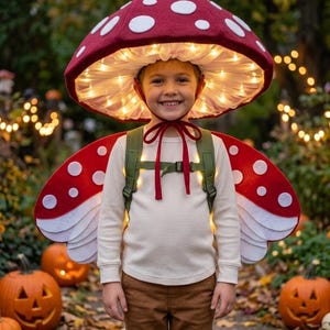 May include: A mushroom costume featuring a large red cap with white spots and integrated warm white string lights, paired with matching red and white spotted wings attached to a green backpack harness, worn over a long-sleeve cream shirt and brown pants.