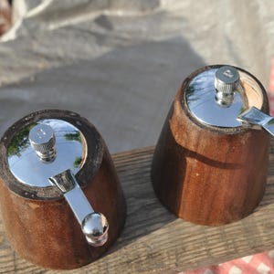 May include: Two wooden pepper grinders with a dark brown finish. Each grinder has a shiny silver top and a silver crank handle. The grinders are set on a wooden surface, with a red and white checkered cloth visible.