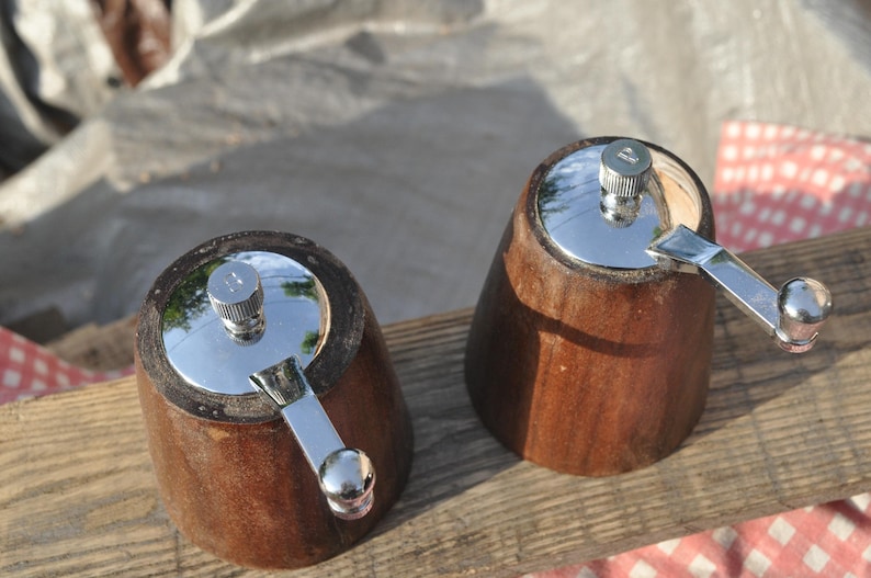 May include: Two wooden pepper grinders with a dark brown finish. Each grinder has a shiny silver top and a silver crank handle. The grinders are set on a wooden surface, with a red and white checkered cloth visible.